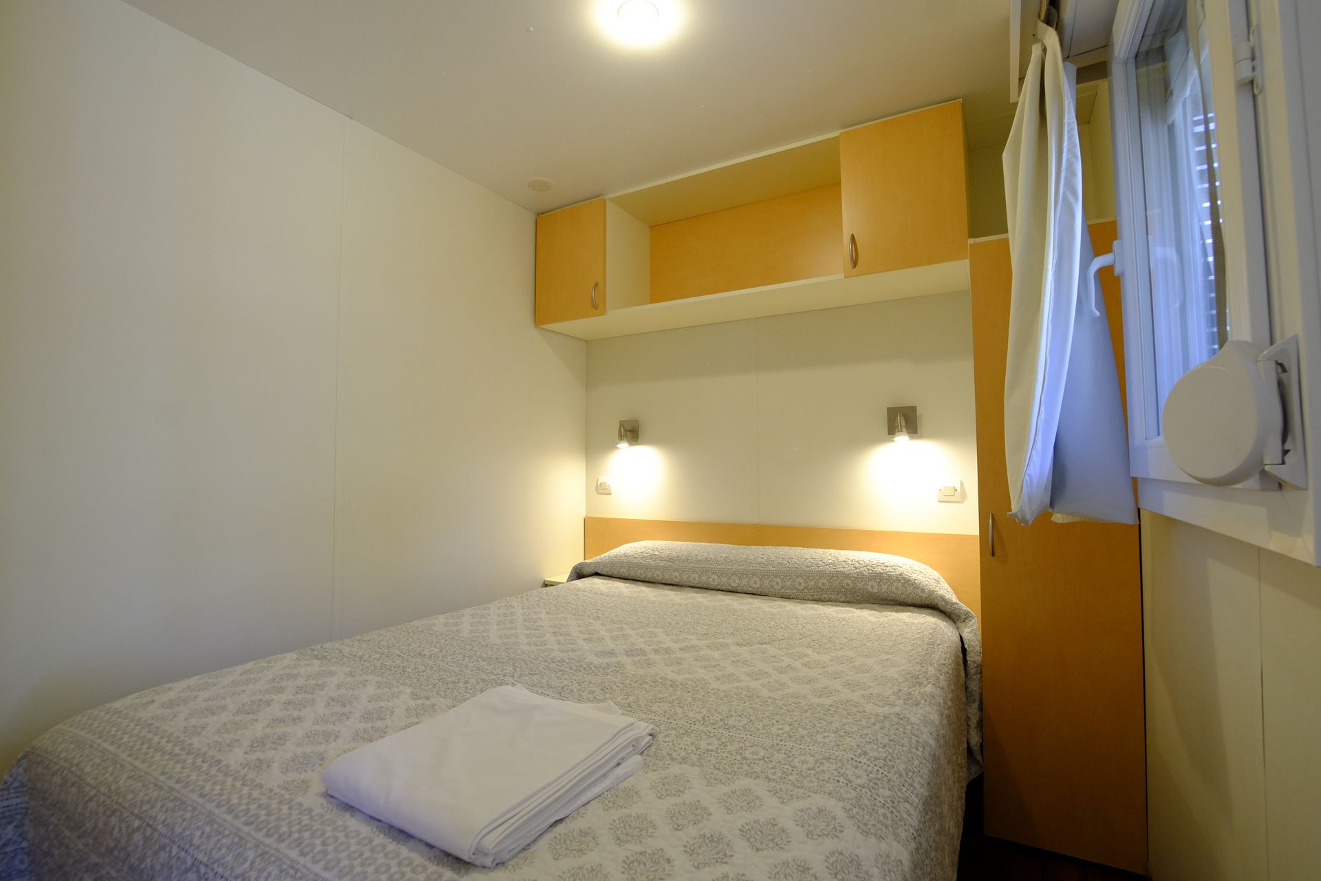 Dormitorio con una cama individual, armarios superiores, lámparas de noche y una pequeña ventana.