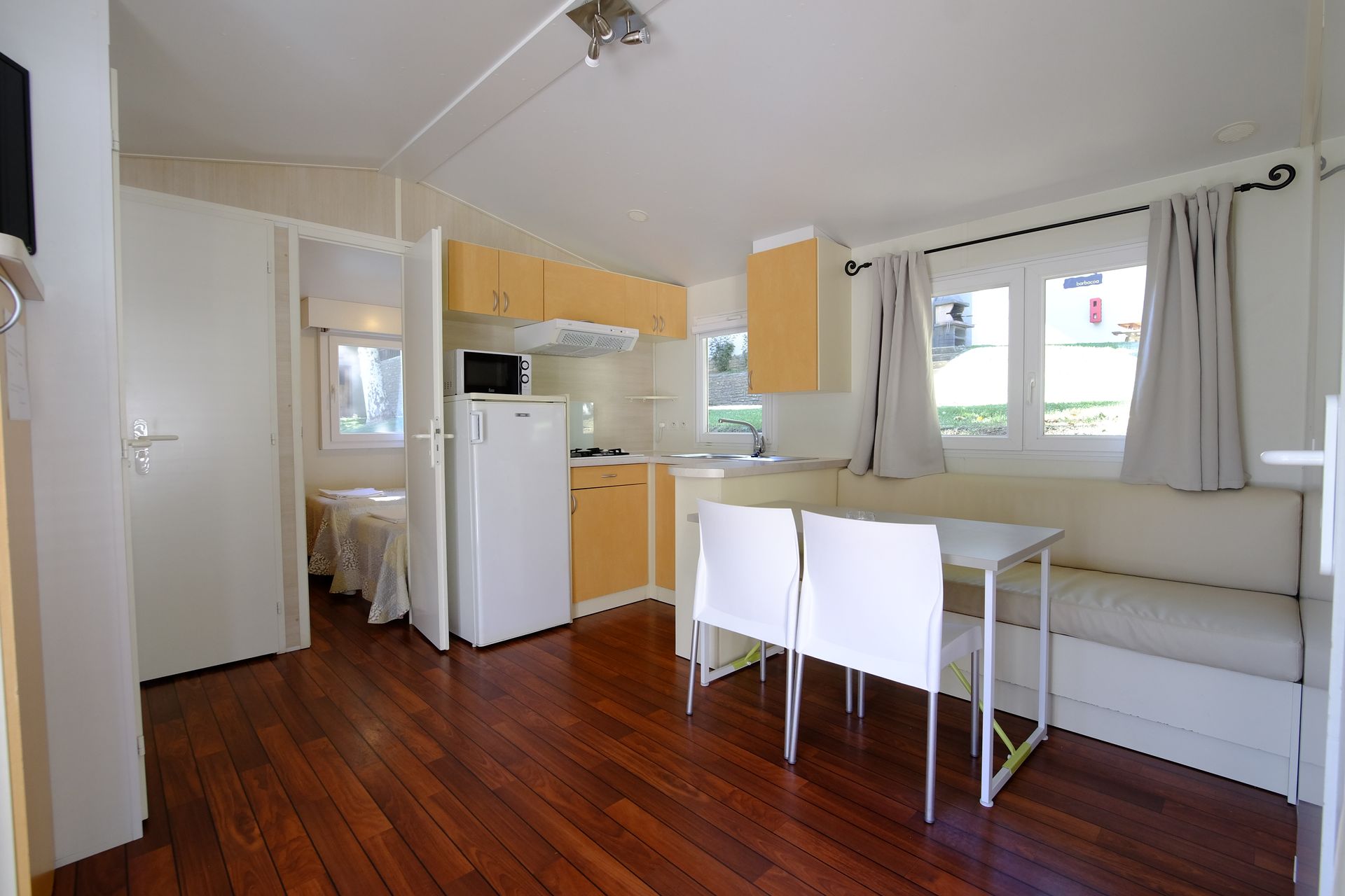 Interior de una pequeña cabaña con cocina, comedor y dormitorio visibles; suelos de madera.
