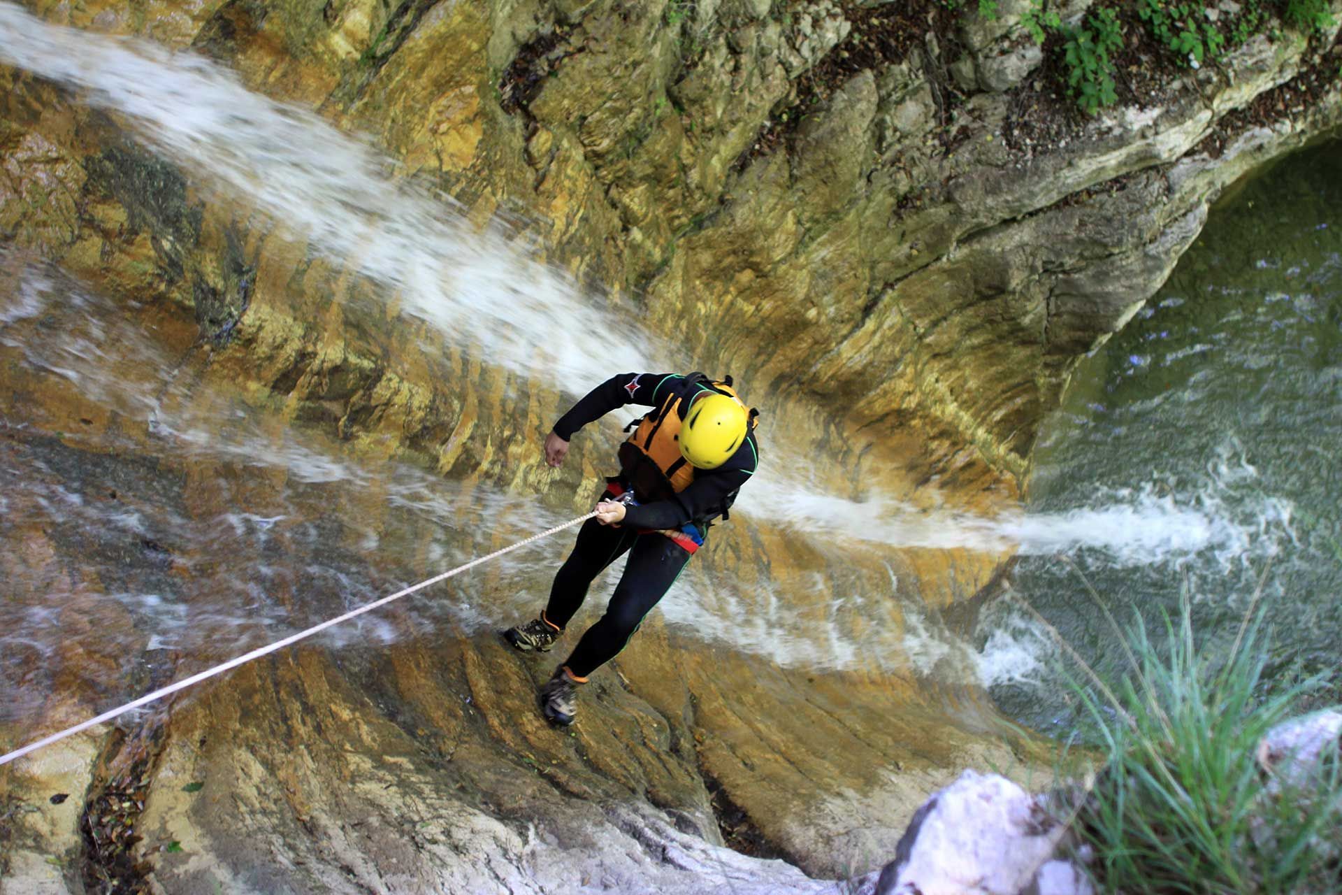 Persona descendiendo en rápel por una cascada, con casco amarillo y traje de neopreno negro. Entorno de cañón rocoso.