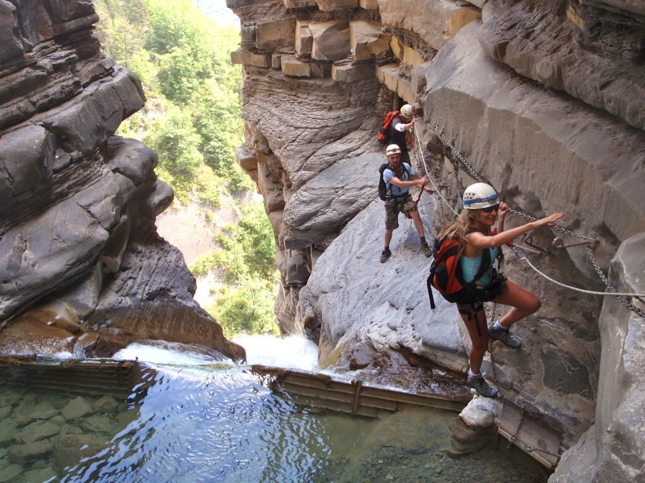 Personas atravesando una estrecha cornisa del cañón, utilizando pasamanos y arneses. Agua cristalina abajo.