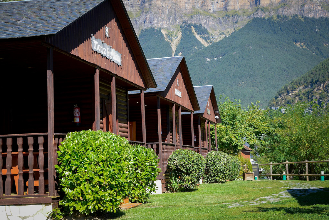 Cabañas de madera con techos oscuros, arbustos verdes y montañas al fondo.