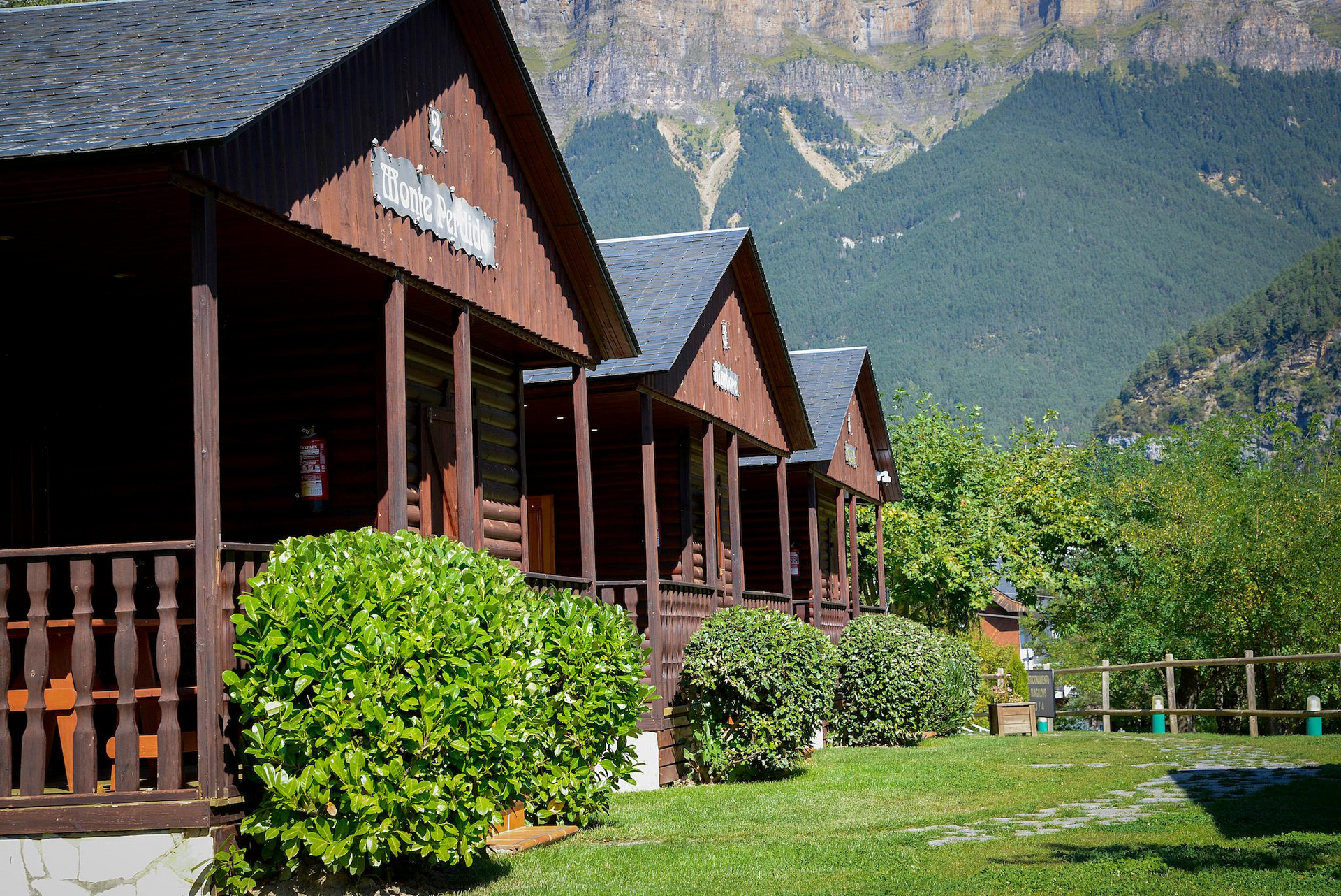Cabañas de madera con techos oscuros, arbustos verdes y montañas al fondo.