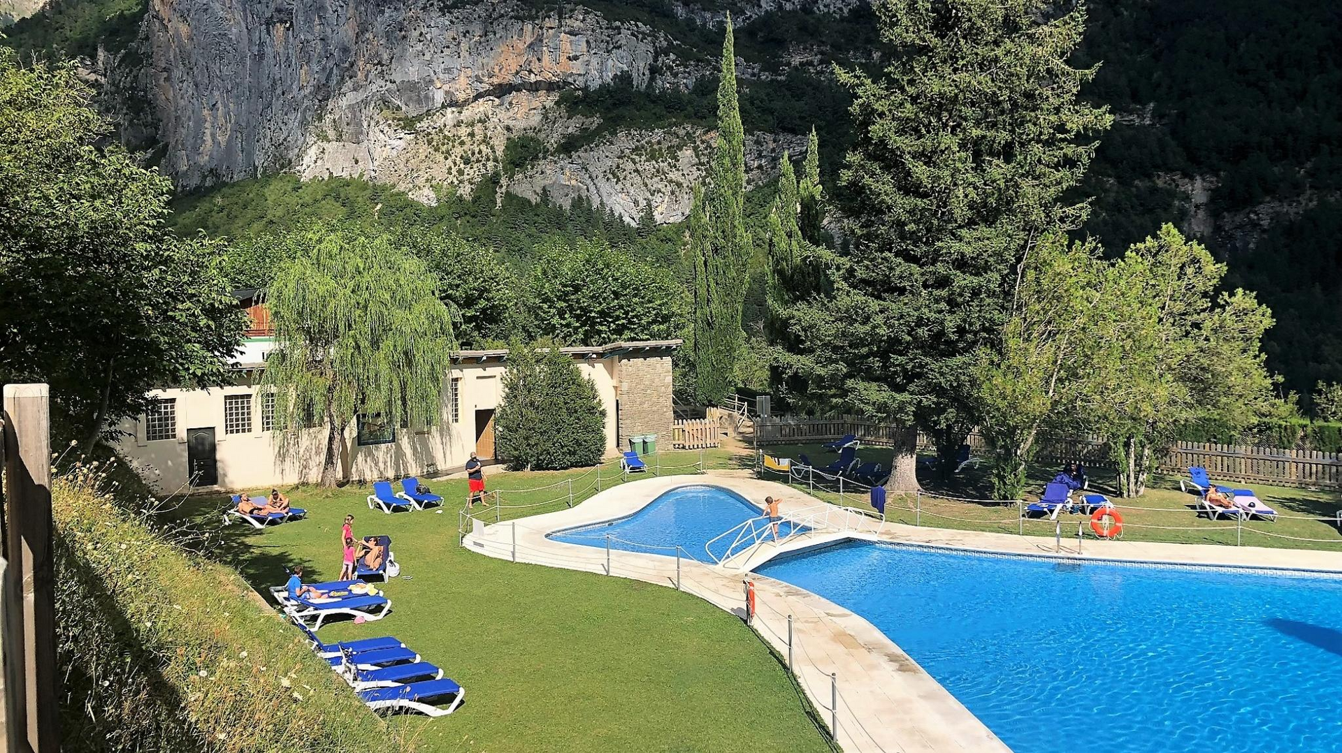 Piscina con gente, césped, árboles y edificios con un acantilado de fondo.