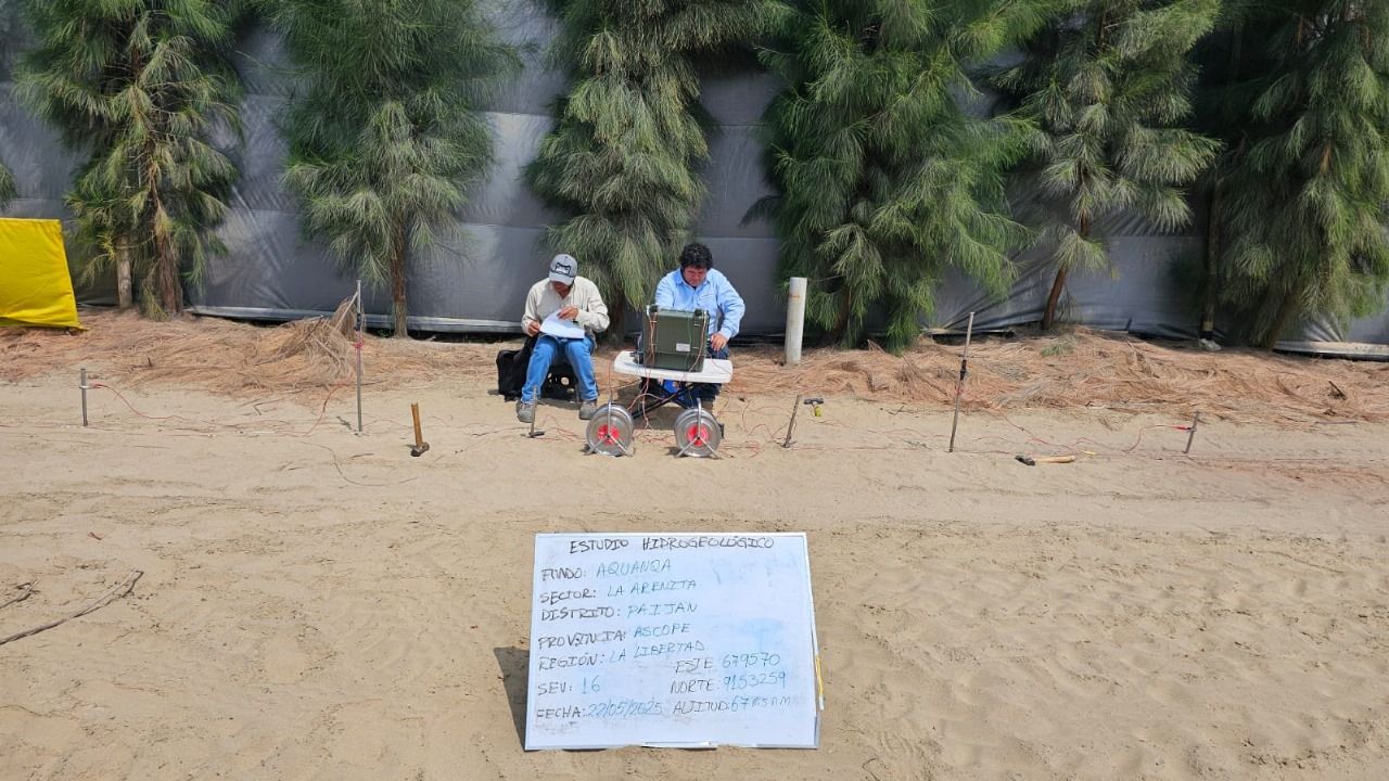 Dos hombres manejando un explorador sobre un terreno arenoso cerca de vegetación y un cartel con texto escrito a mano.