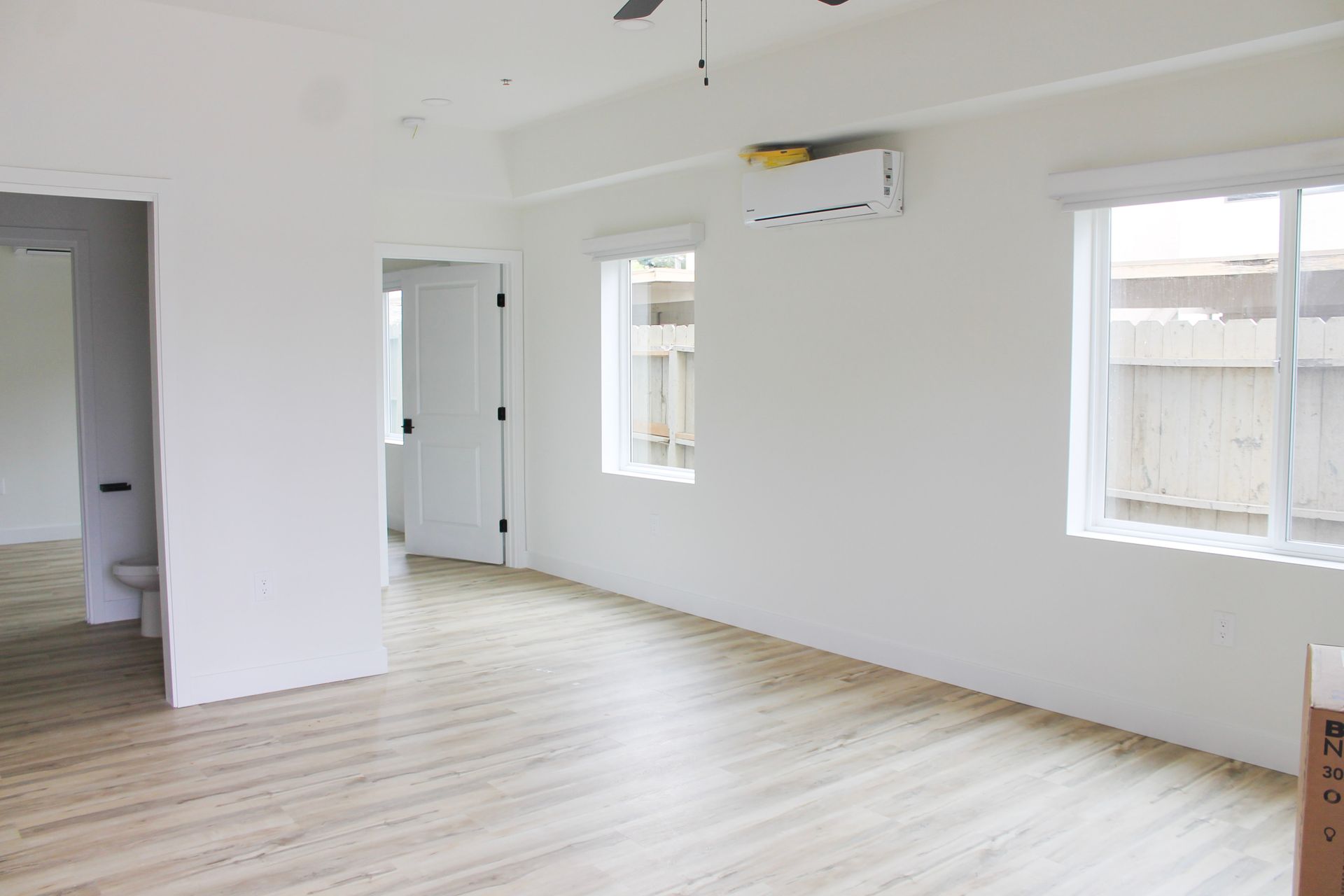 Empty white room with wood-look floors, two windows, doorway, and a wall-mounted air conditioner.