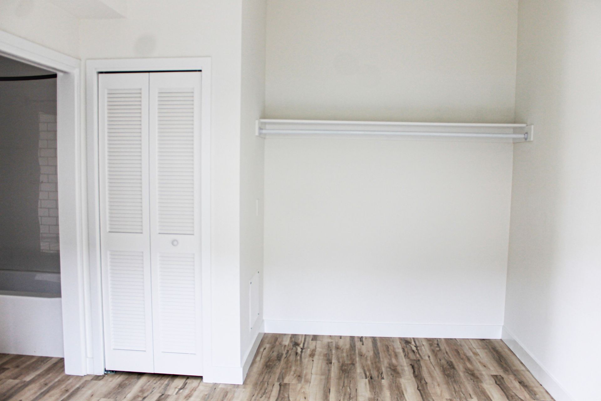 Empty white closet and folding door in room with wood-look floor; door to bathroom visible.