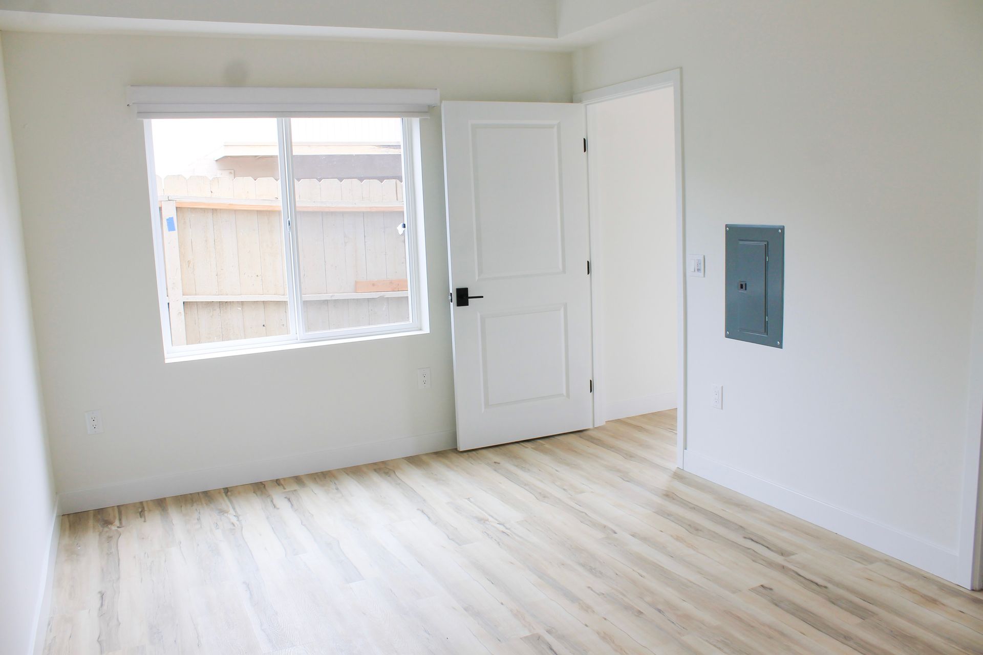 Empty room with light wood floors, white walls, and a window. A door and electrical box are also visible.