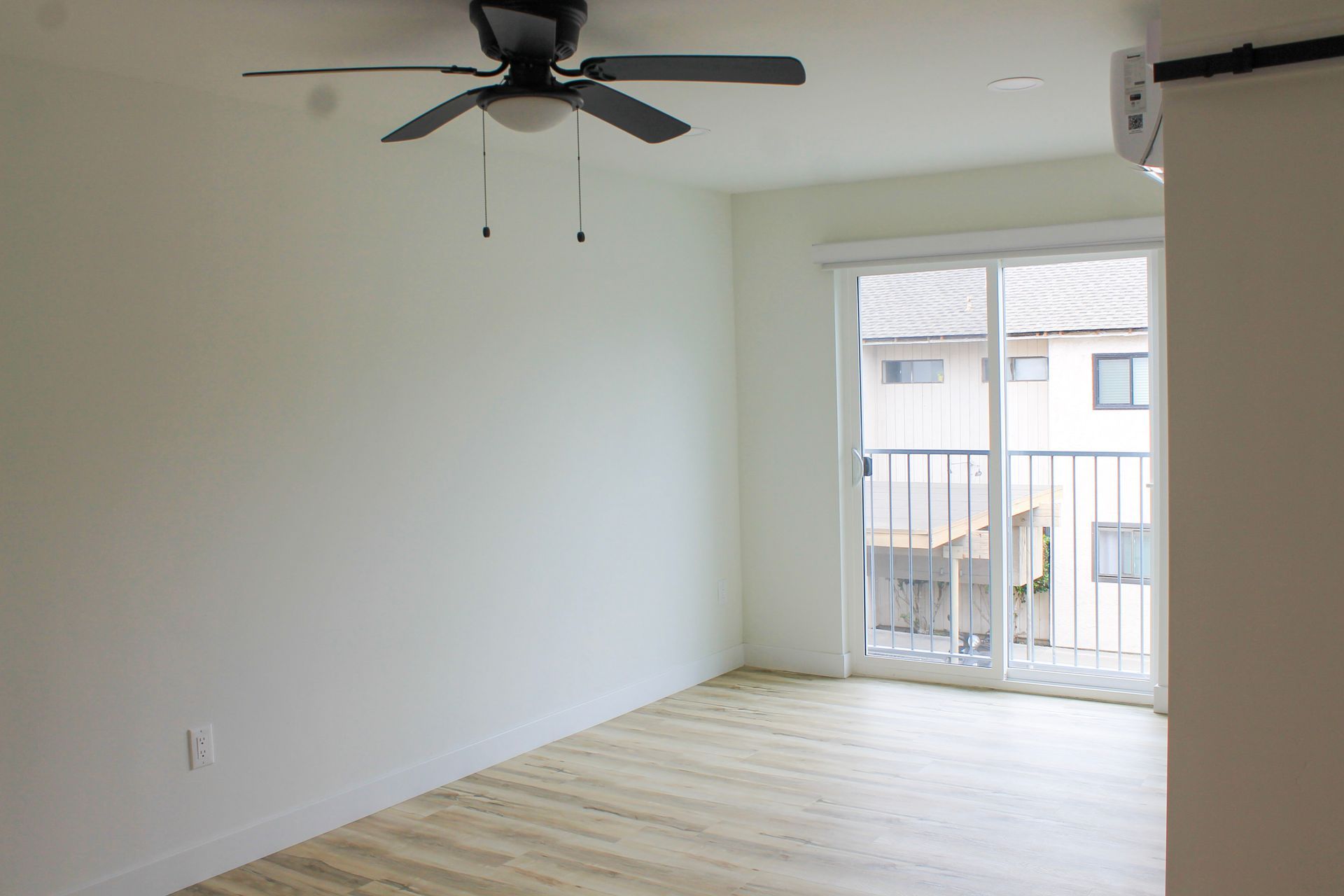 Empty room with light wood floor, sliding glass door to balcony, ceiling fan.
