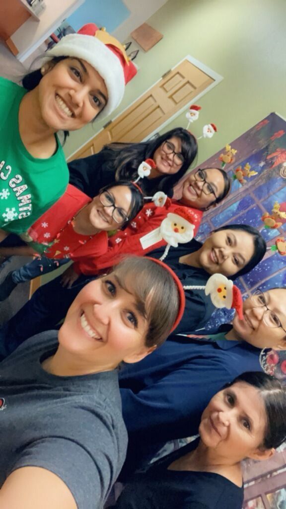 Group of smiling people in holiday attire, posing for a selfie. Festive decorations in background.