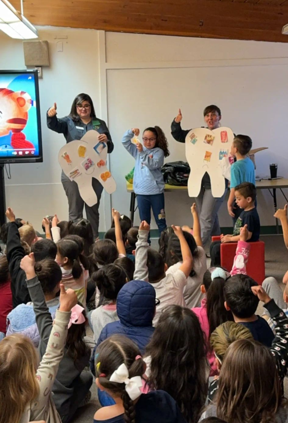 Children in a classroom with instructors holding tooth cutouts; raised hands, smiling faces, learning about dental hygiene.