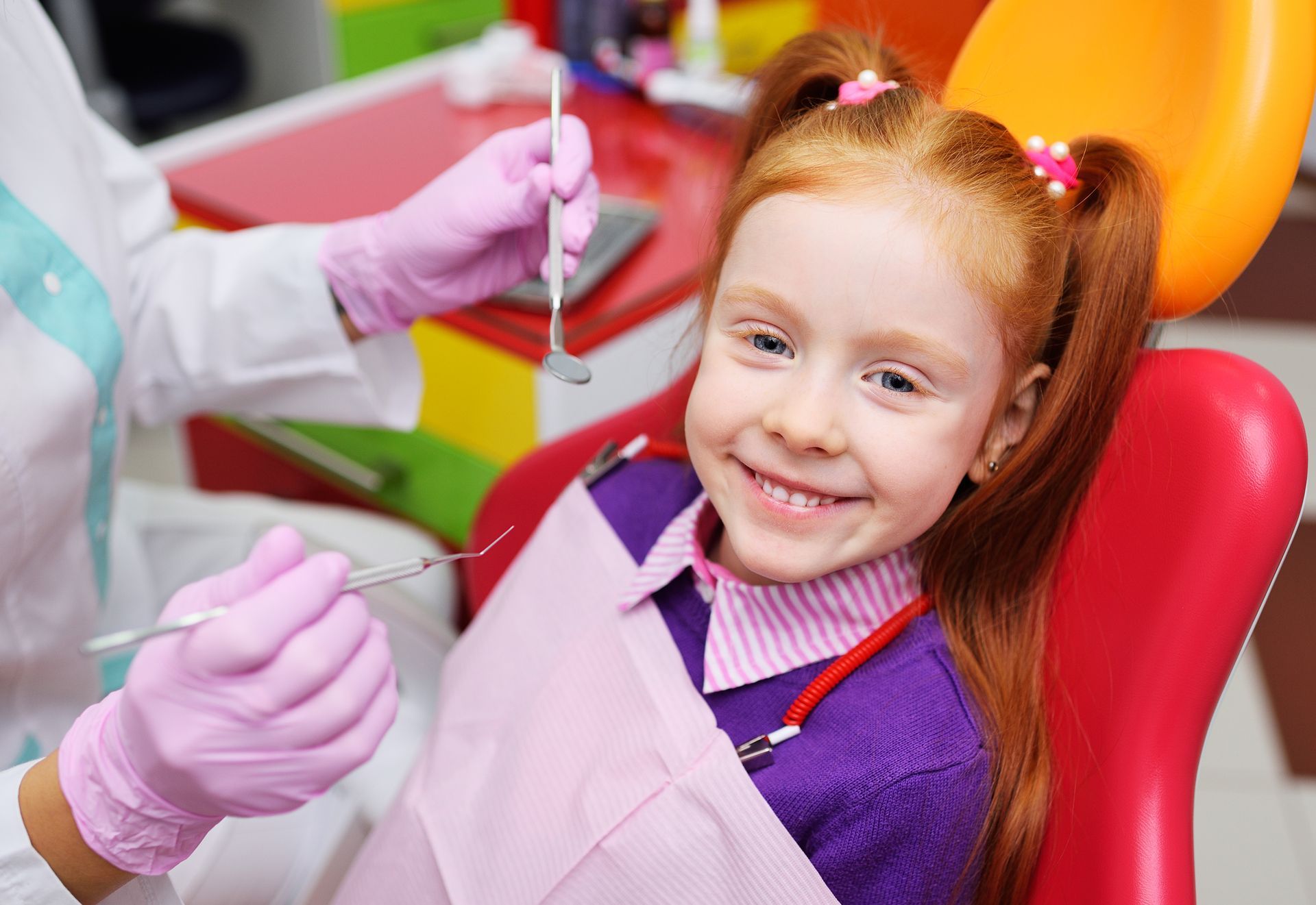 A red-haired girl smiles in a dentist's chair. A dentist in pink gloves holds tools, ready for an exam.