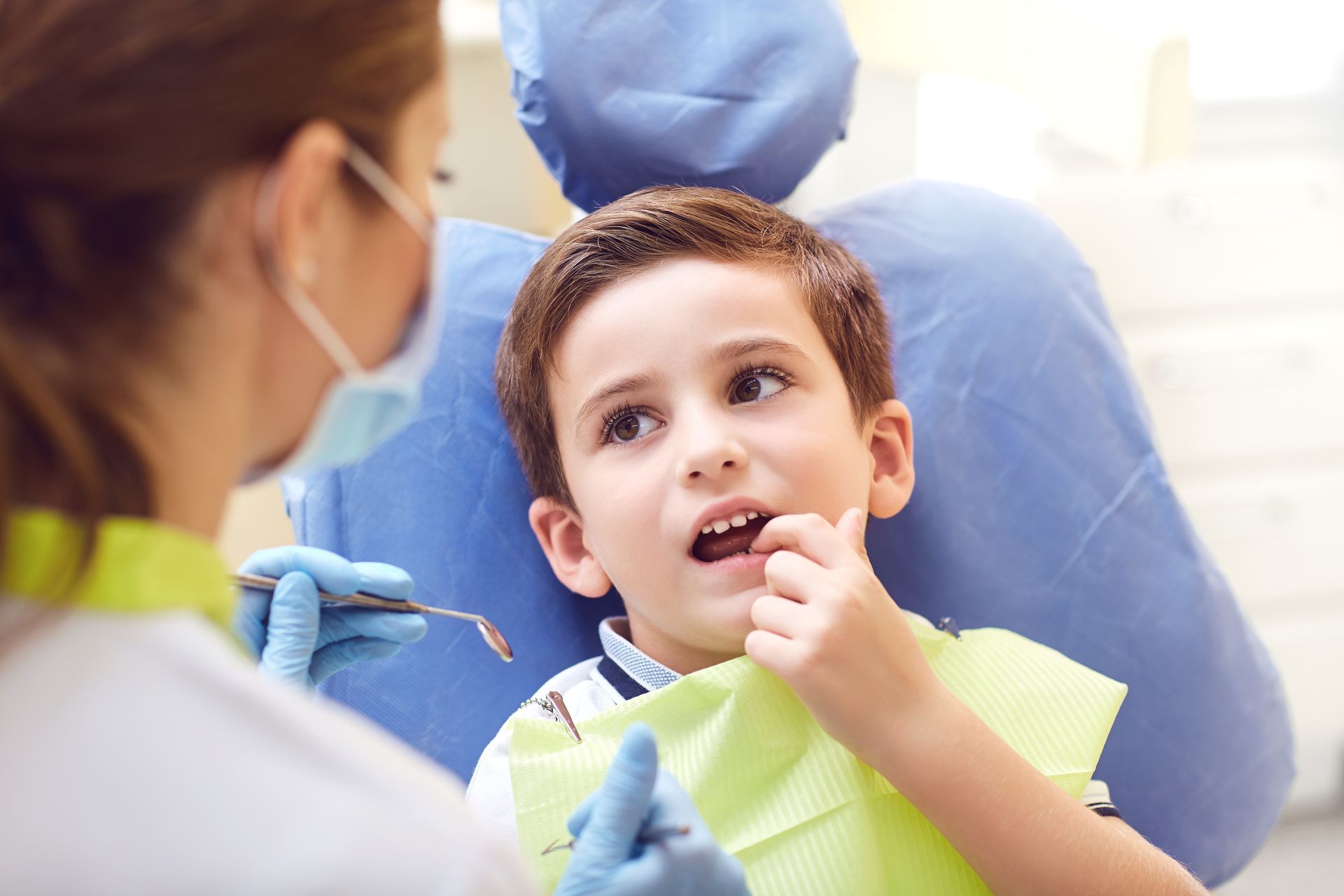 Boy at dentist, pointing to mouth, examining tooth with dentist in blue gloves and mask.