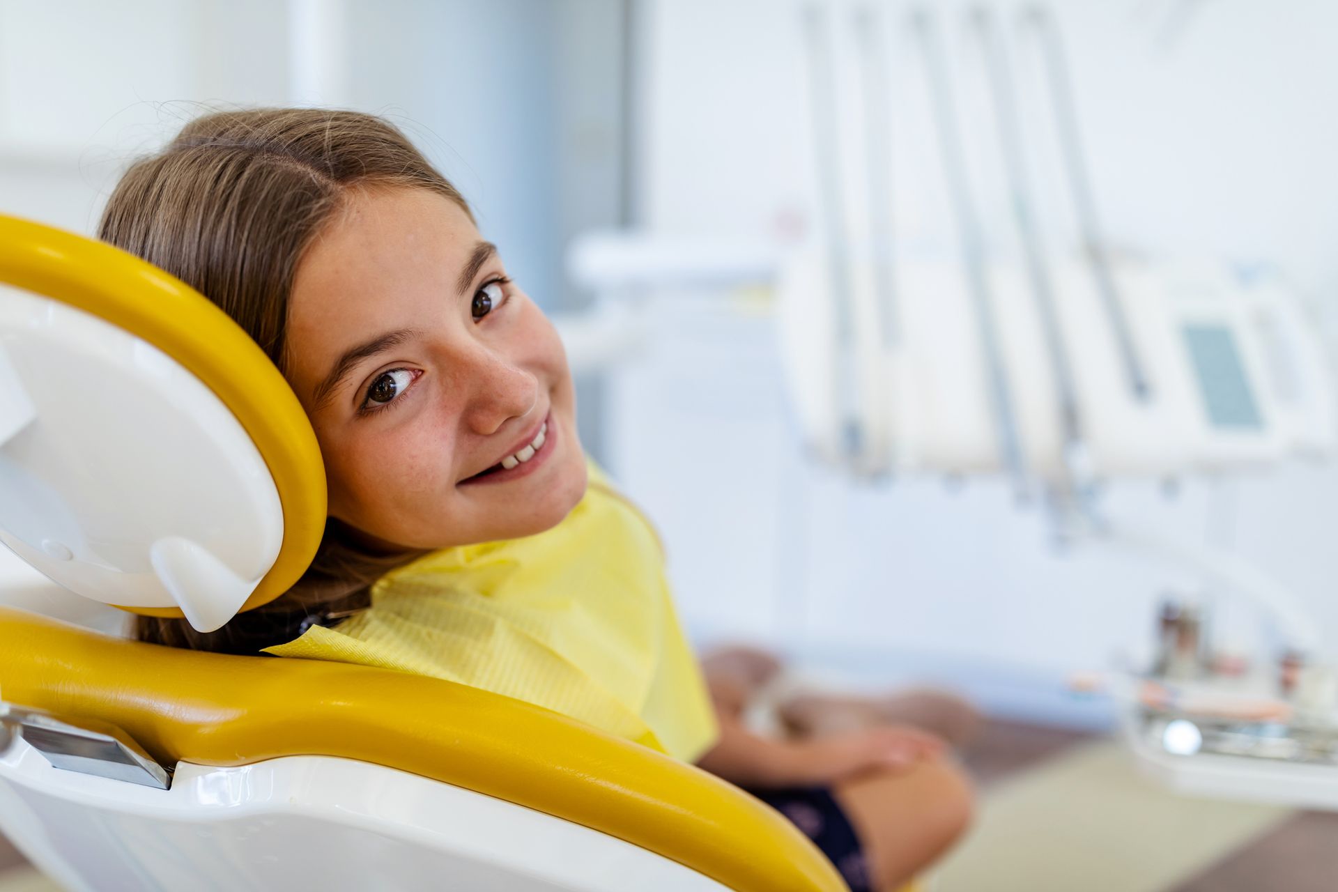 Girl in a dental chair, smiling, wearing yellow, with dental tools in the background.