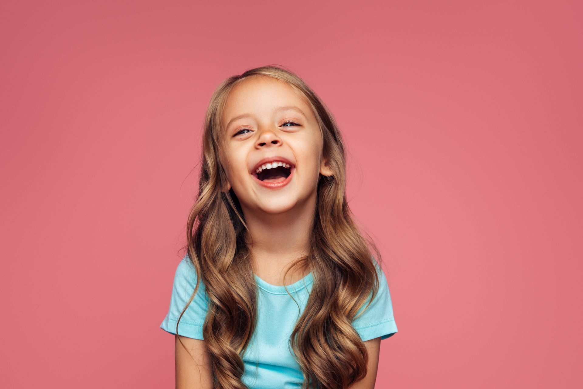 Girl with long wavy brown hair laughs, wearing a blue shirt, set against a pink background.