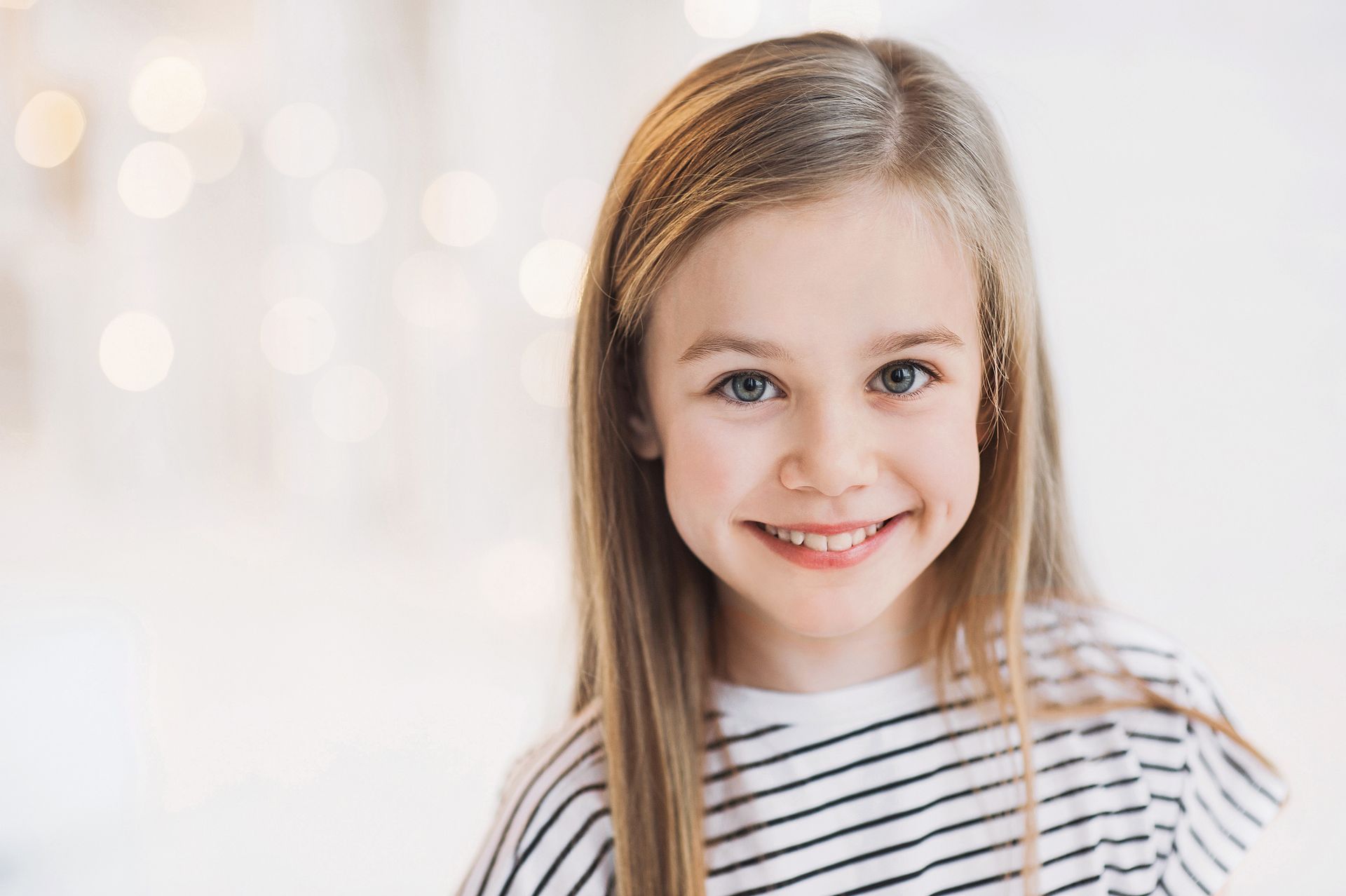 Blonde girl with a striped shirt smiles at the camera, soft background lights.