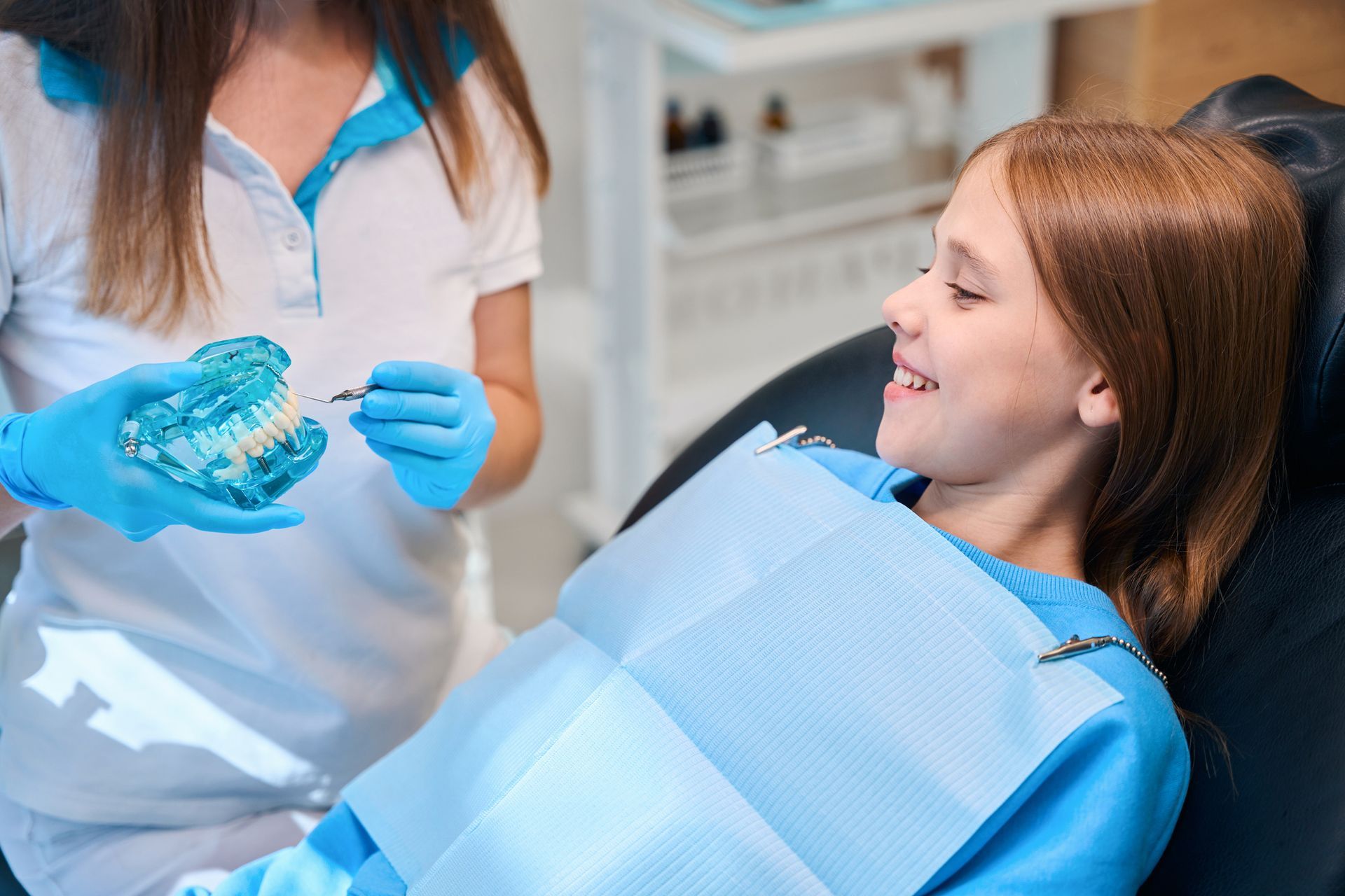 A dentist showing a teeth model to a smiling girl in a dental chair, with blue gloves and a bib.