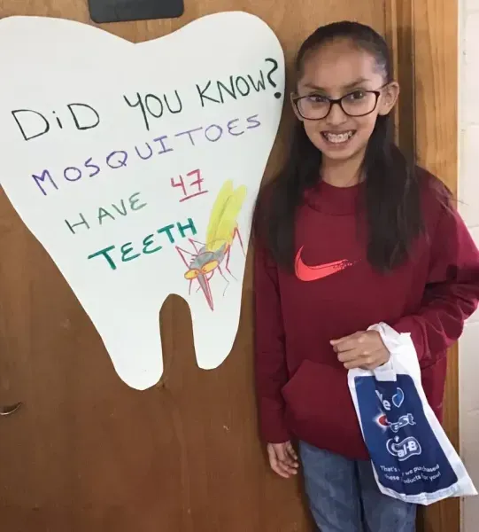 Girl with glasses holding a bag stands next to a tooth-shaped sign stating