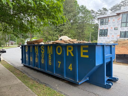 Blue dumpster on the side of a road, filled with debris, near a building under construction. 