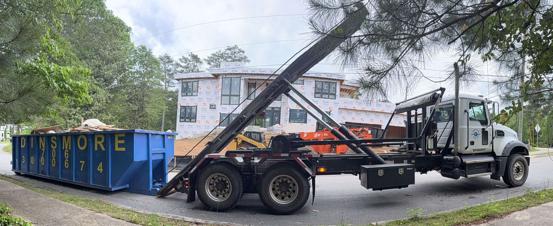 A blue dumpster is being lifted by a truck in front of a partially constructed building, trees surrounding.