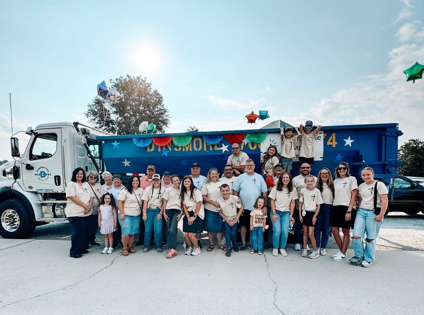 Group of people pose with a blue dump truck, outdoors on a sunny day. Some wear matching shirts.