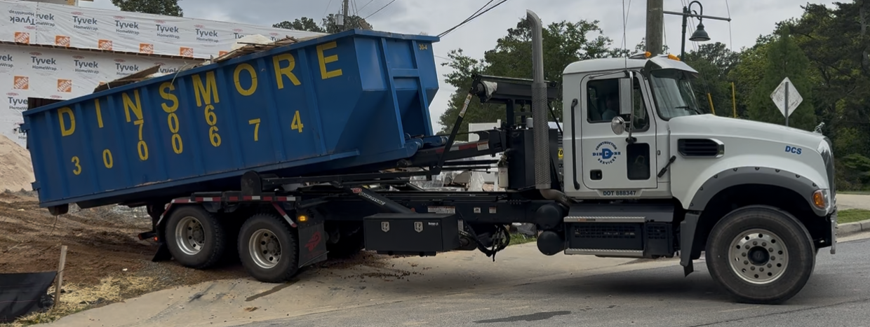 A white Dinsmore truck with a blue dumpster parked on a street near a construction site.