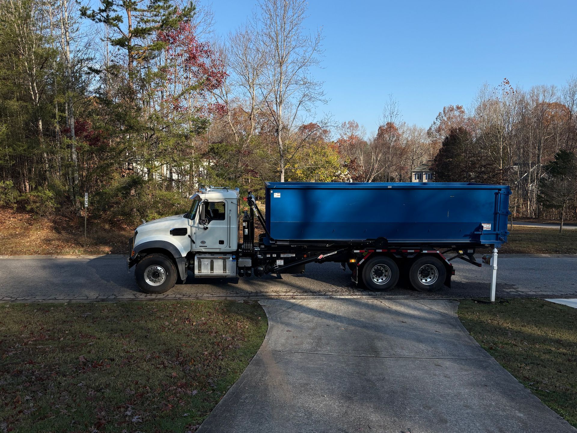 A white truck with a blue dumpster on a driveway, set in a residential area with trees.