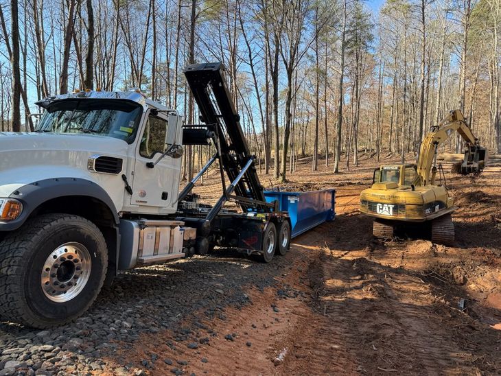 A white dump truck with a blue container next to a yellow excavator in a wooded area.