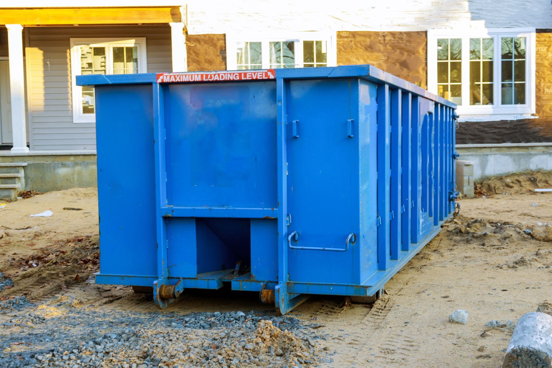 Blue construction dumpster on a dirt lot in front of a house under construction.