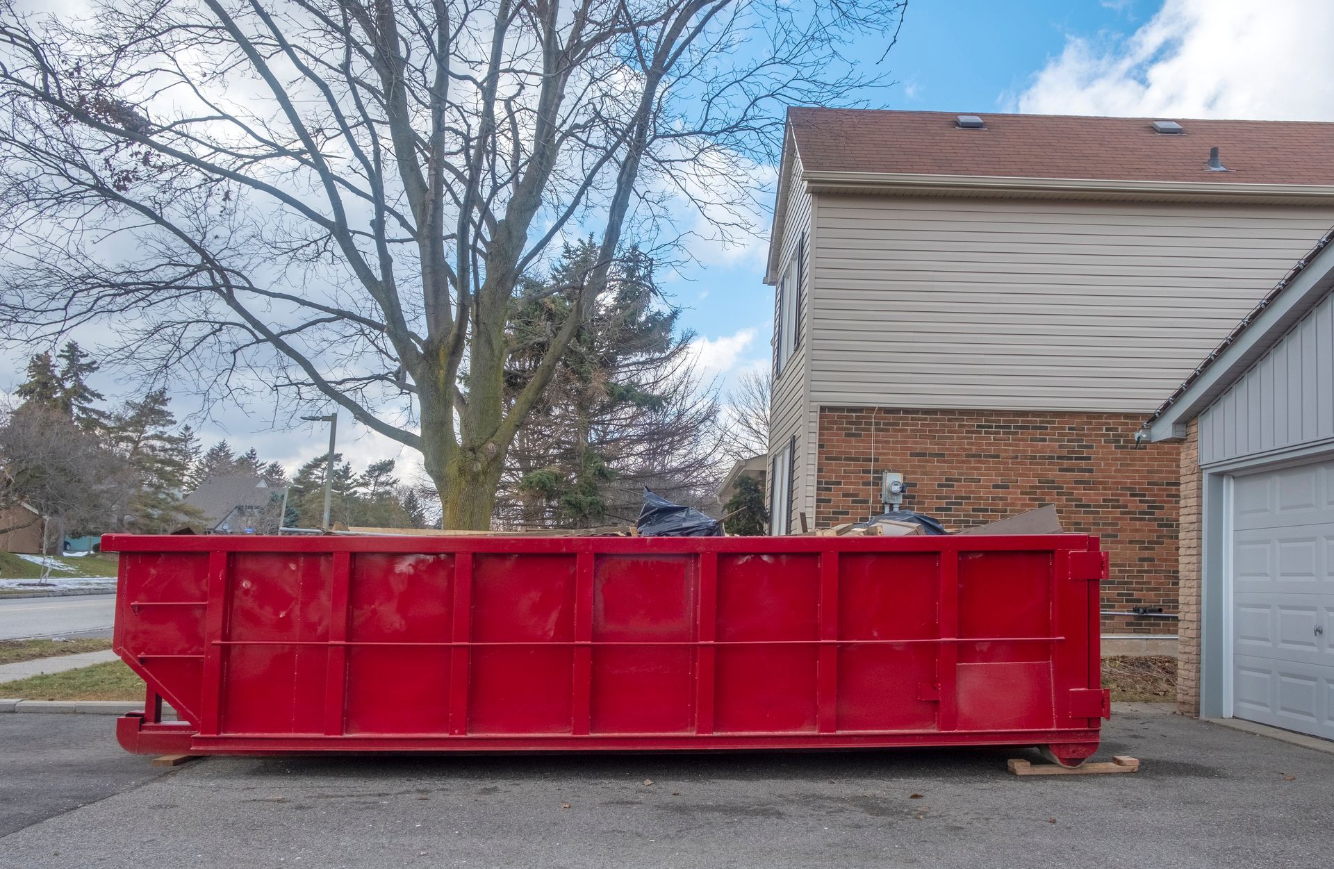 Large red dumpster on a paved driveway next to a building and tree.