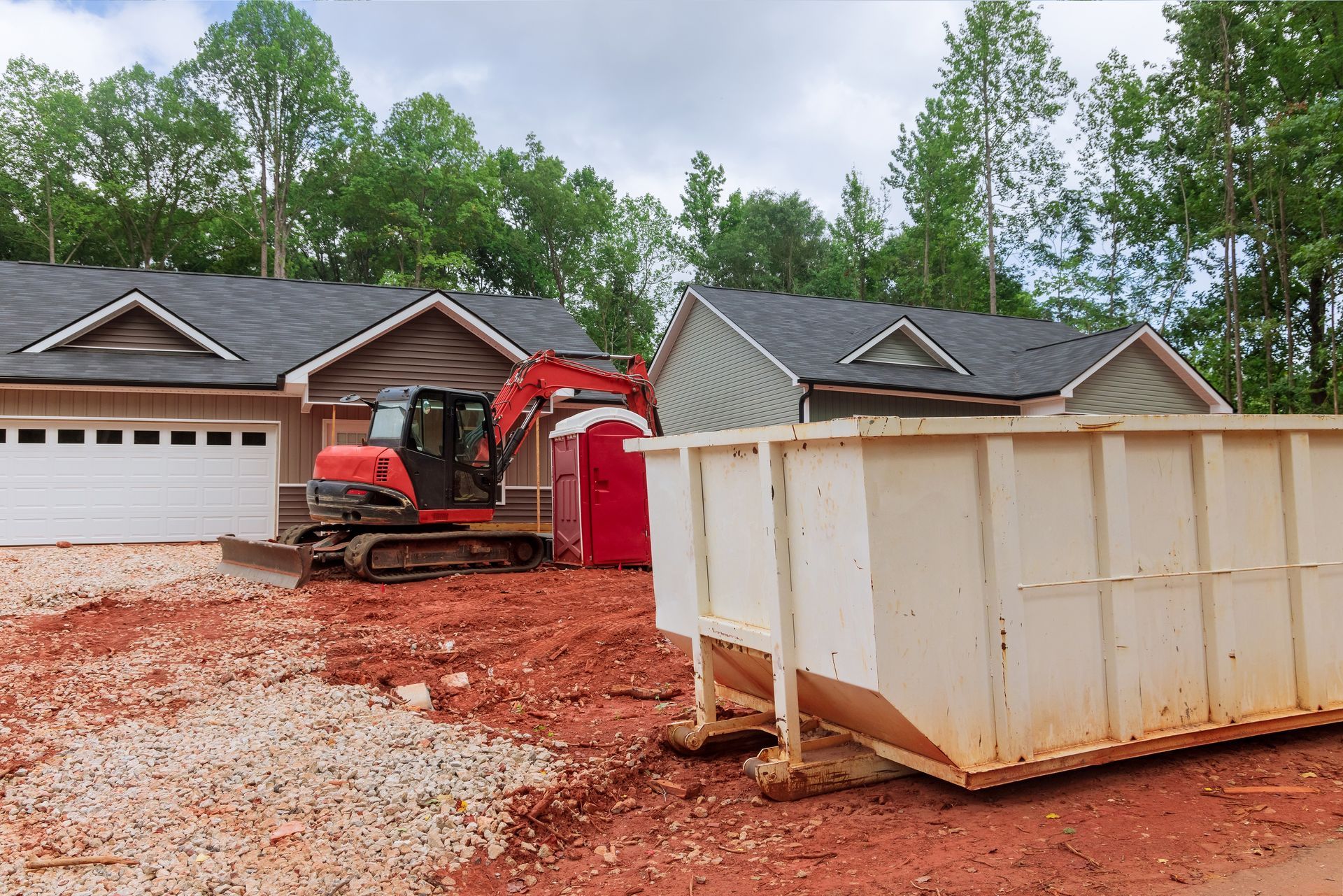 Construction site with red excavator, portable toilet, and dumpster near new houses.
