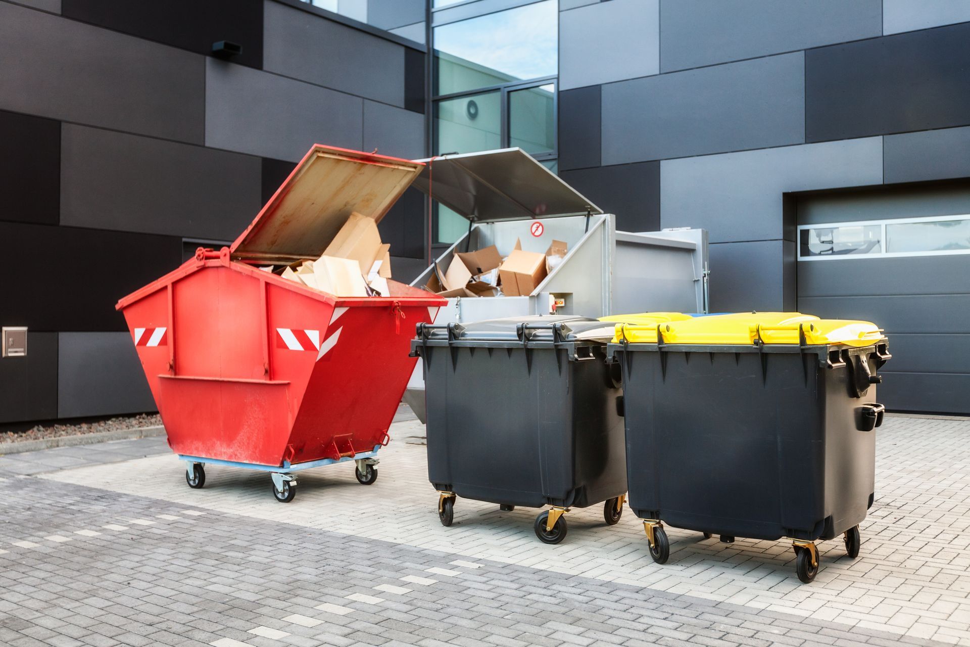Red, black, and yellow dumpsters with lids open and closed, set against a dark building and brick pavement.