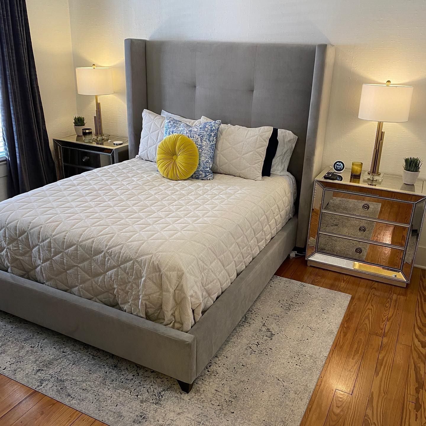 A gray upholstered bed with a tall headboard, mirrored nightstands, and a white bedspread in a bedroom.