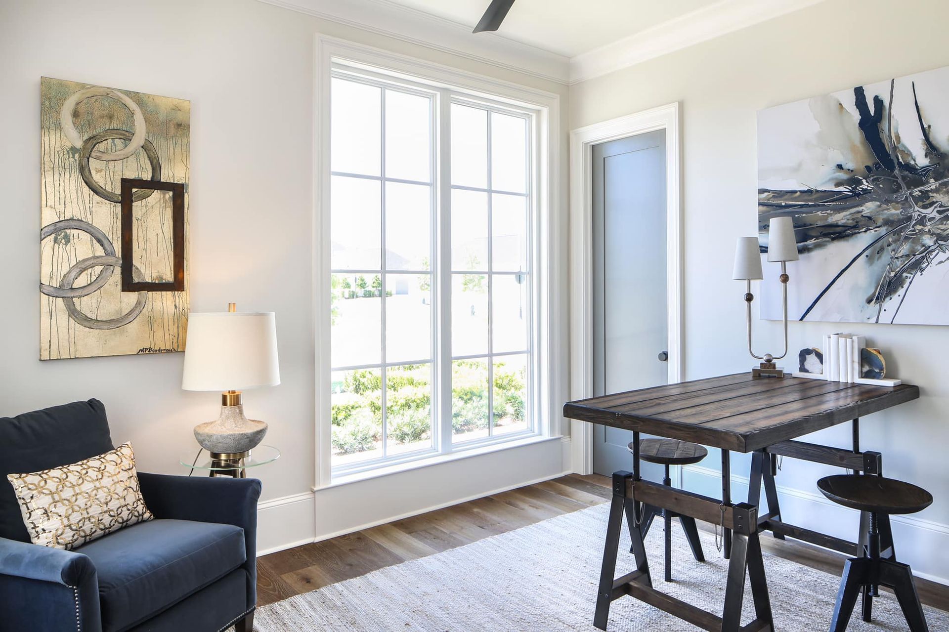 Home office with blue armchair, dark wood desk, large window, artwork, and rug.
