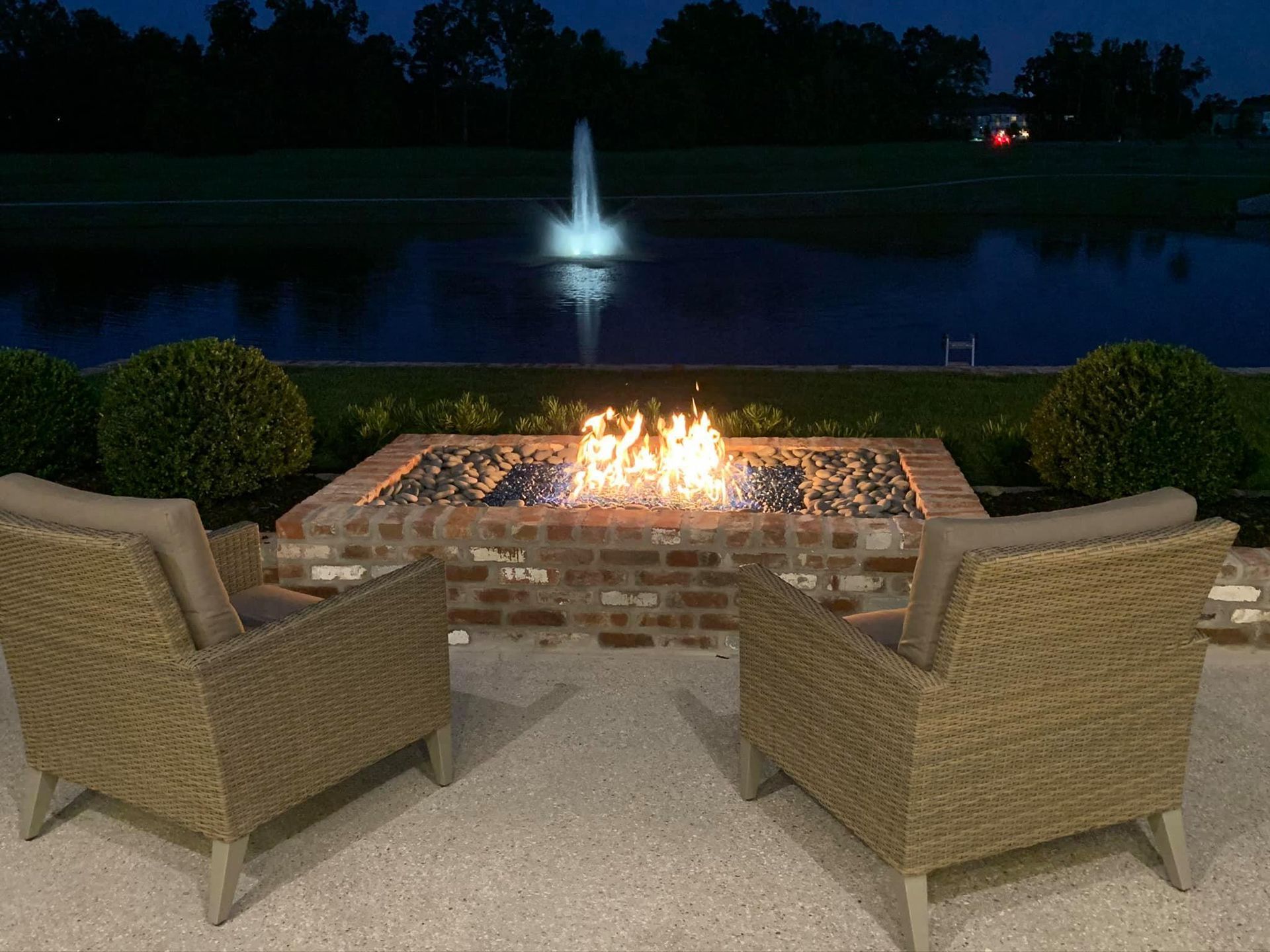 Two wicker chairs face a brick fire pit with a water fountain in the background at dusk.