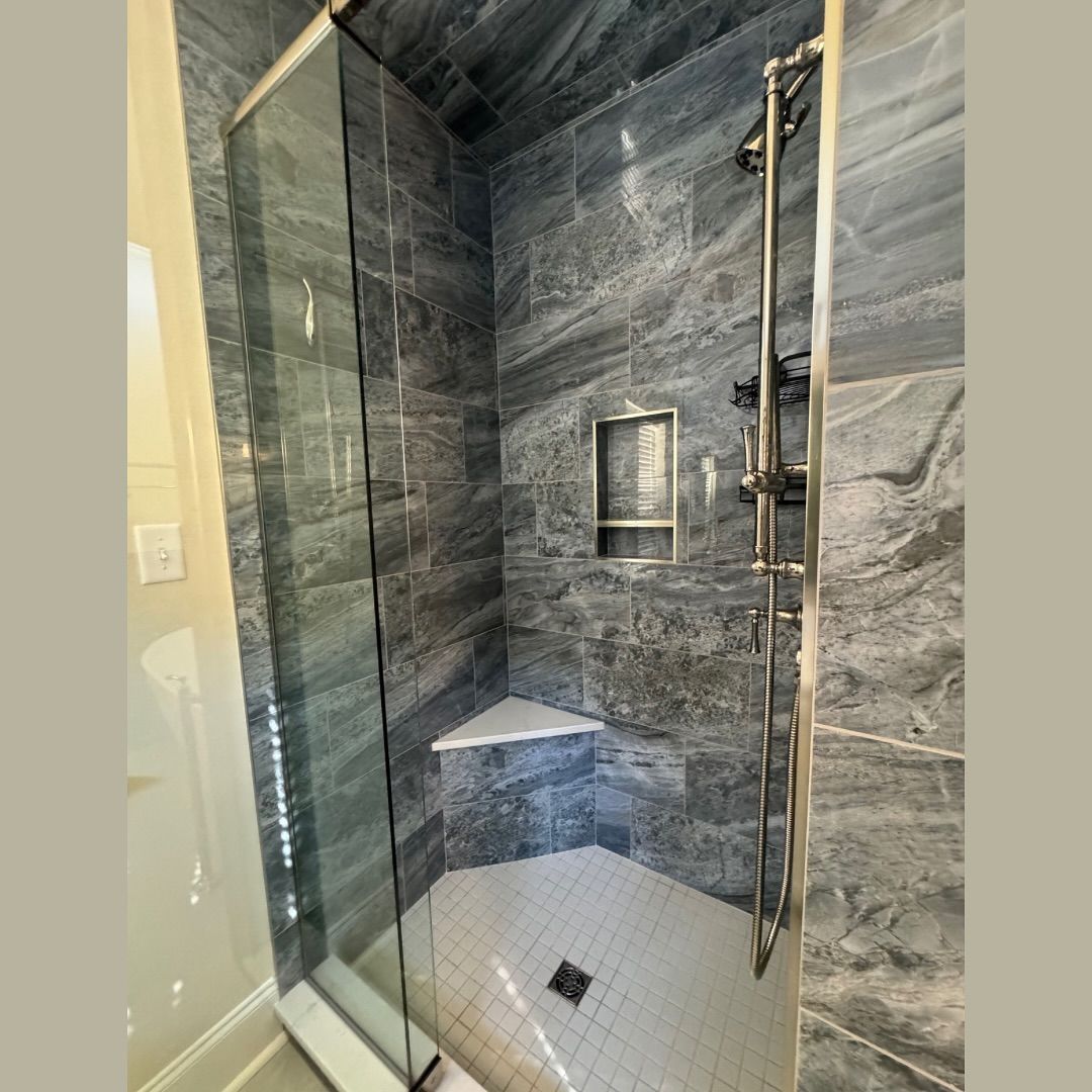 Shower with gray marble-look tile walls, glass door, corner shelf, and white hexagonal floor tiles.