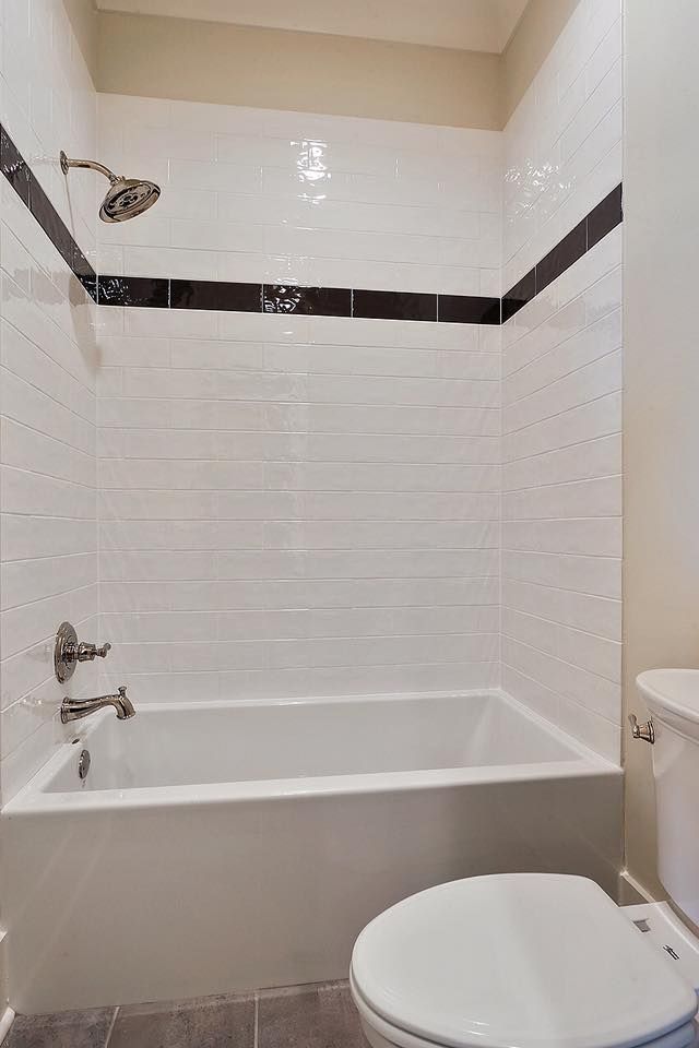 White-tiled bathroom with tub, showerhead, dark stripe, and toilet.
