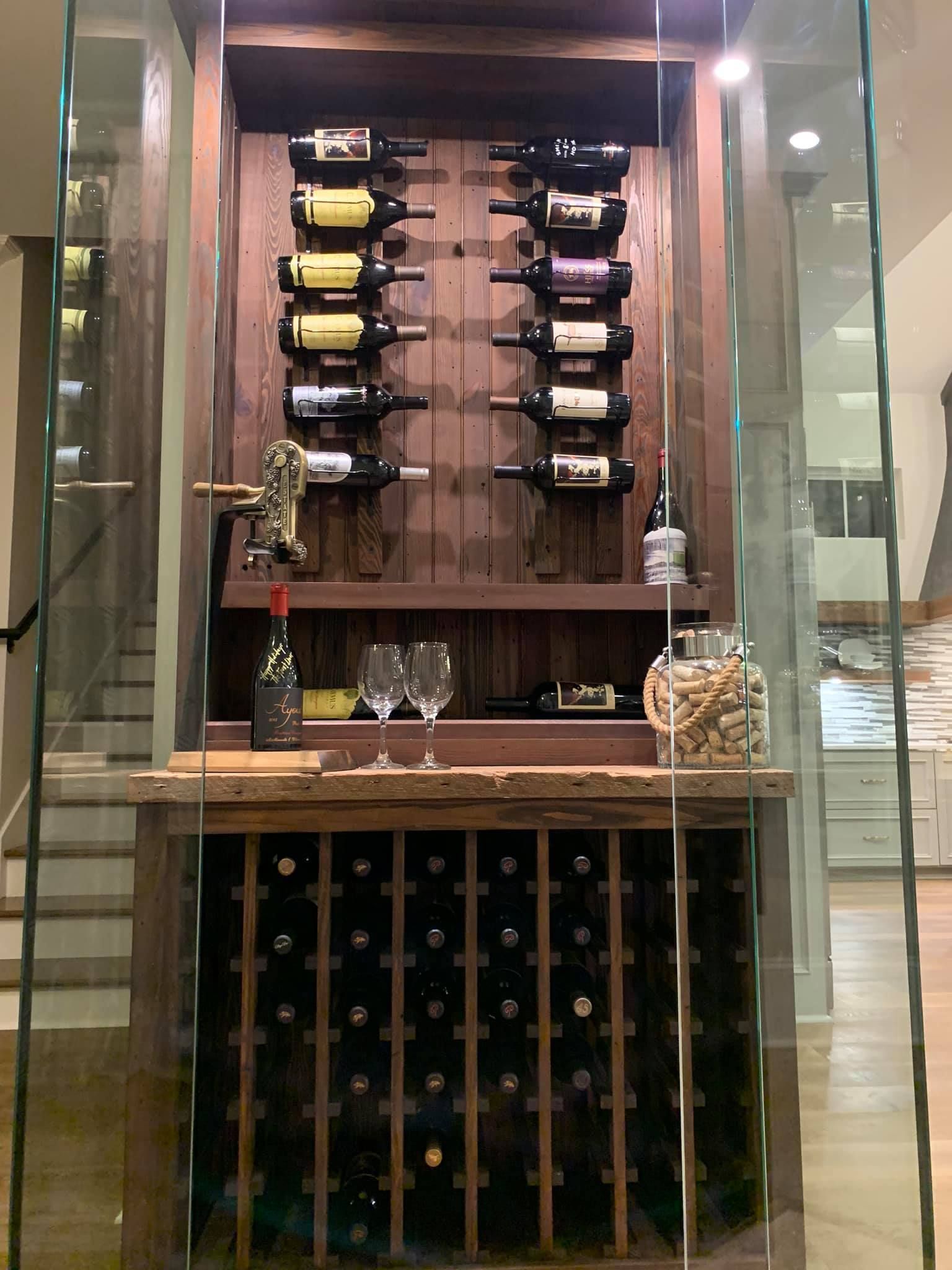 Wine cellar with wooden shelving, holding bottles. Enclosed in glass with stairs visible.