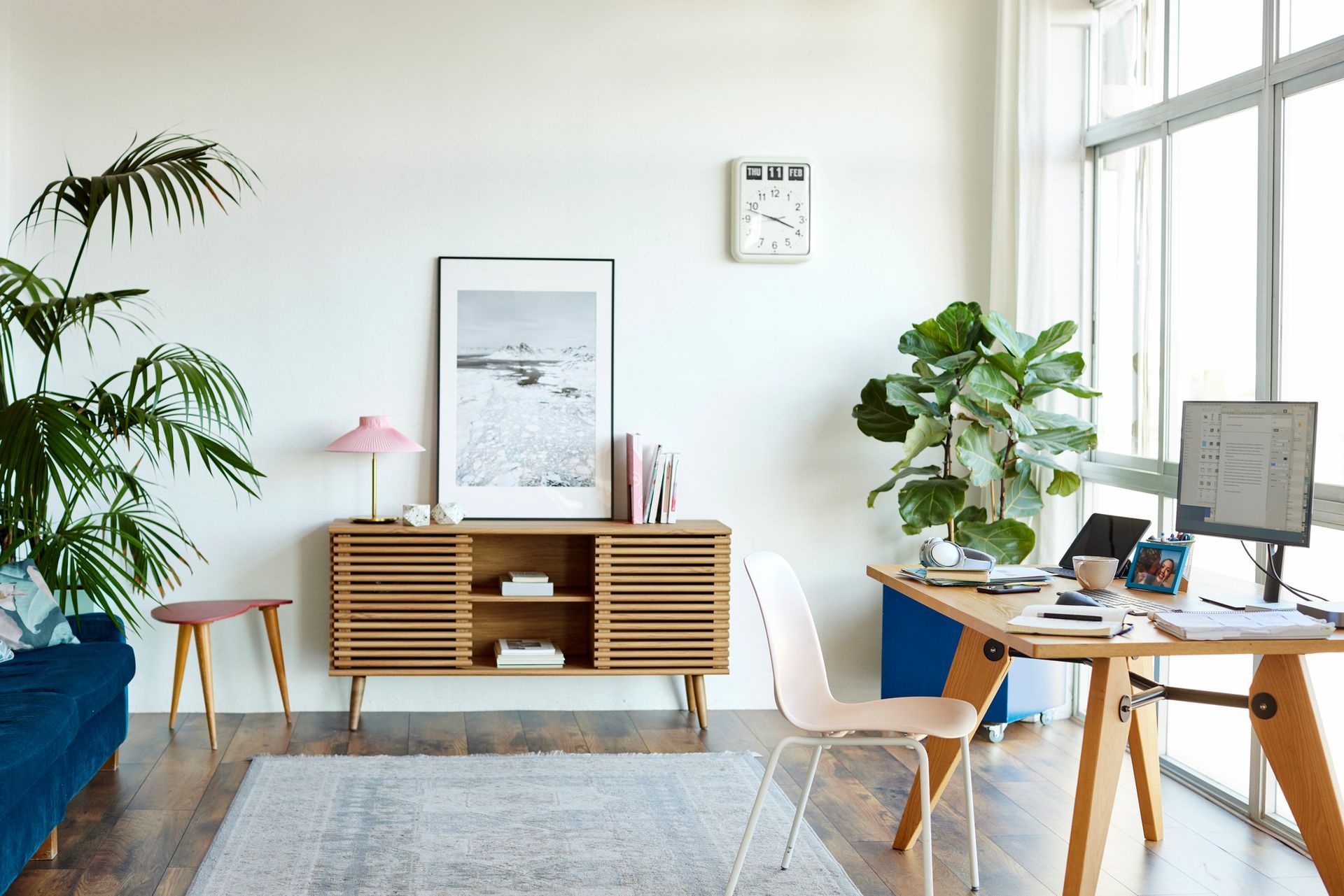 A well-lit living room with a desk, cabinet, plants, and large window; a desk with a laptop, a blue couch.