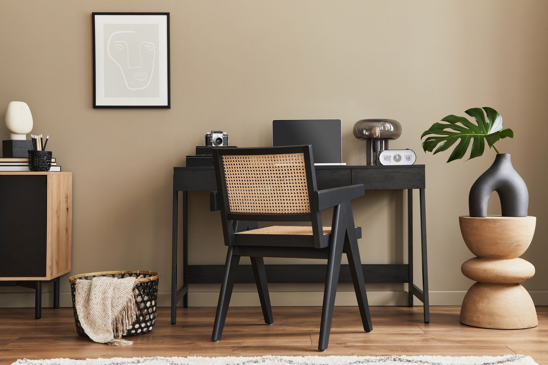 A stylish home office with a black desk, rattan chair, and decor, with a muted brown wall.