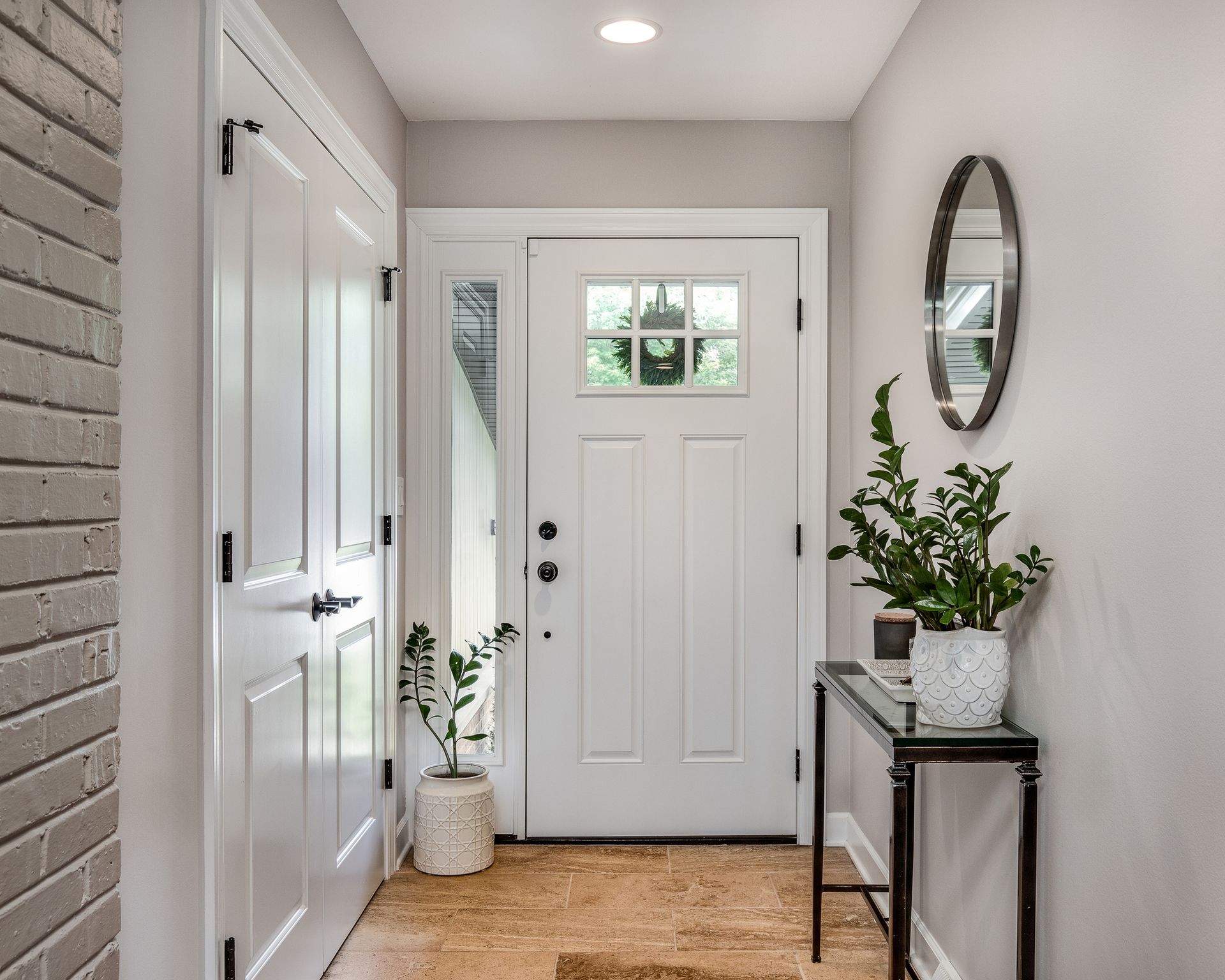 White entryway with door, console table, plants, and oval mirror.