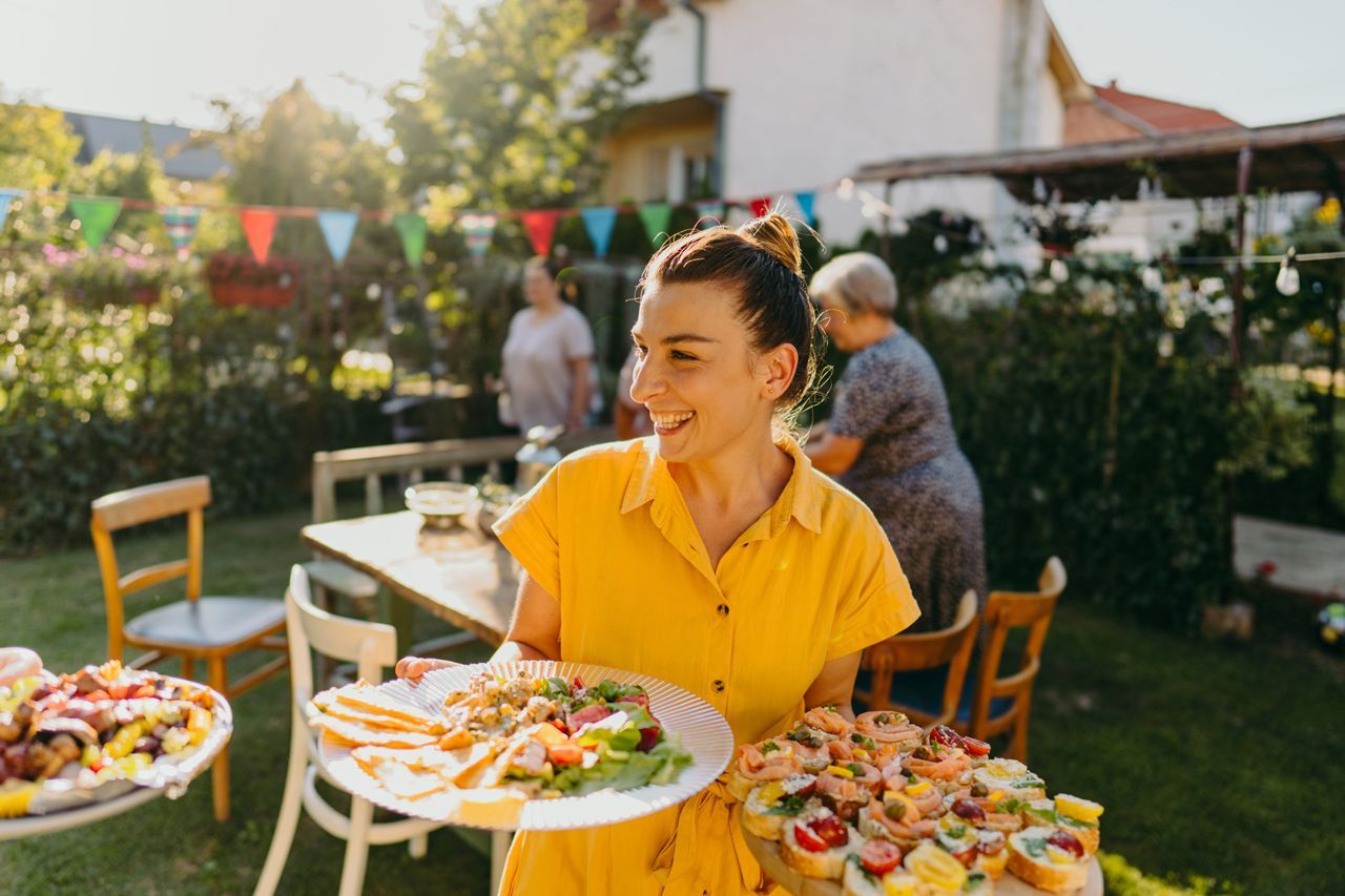 A woman is holding plates of food at a backyard party.