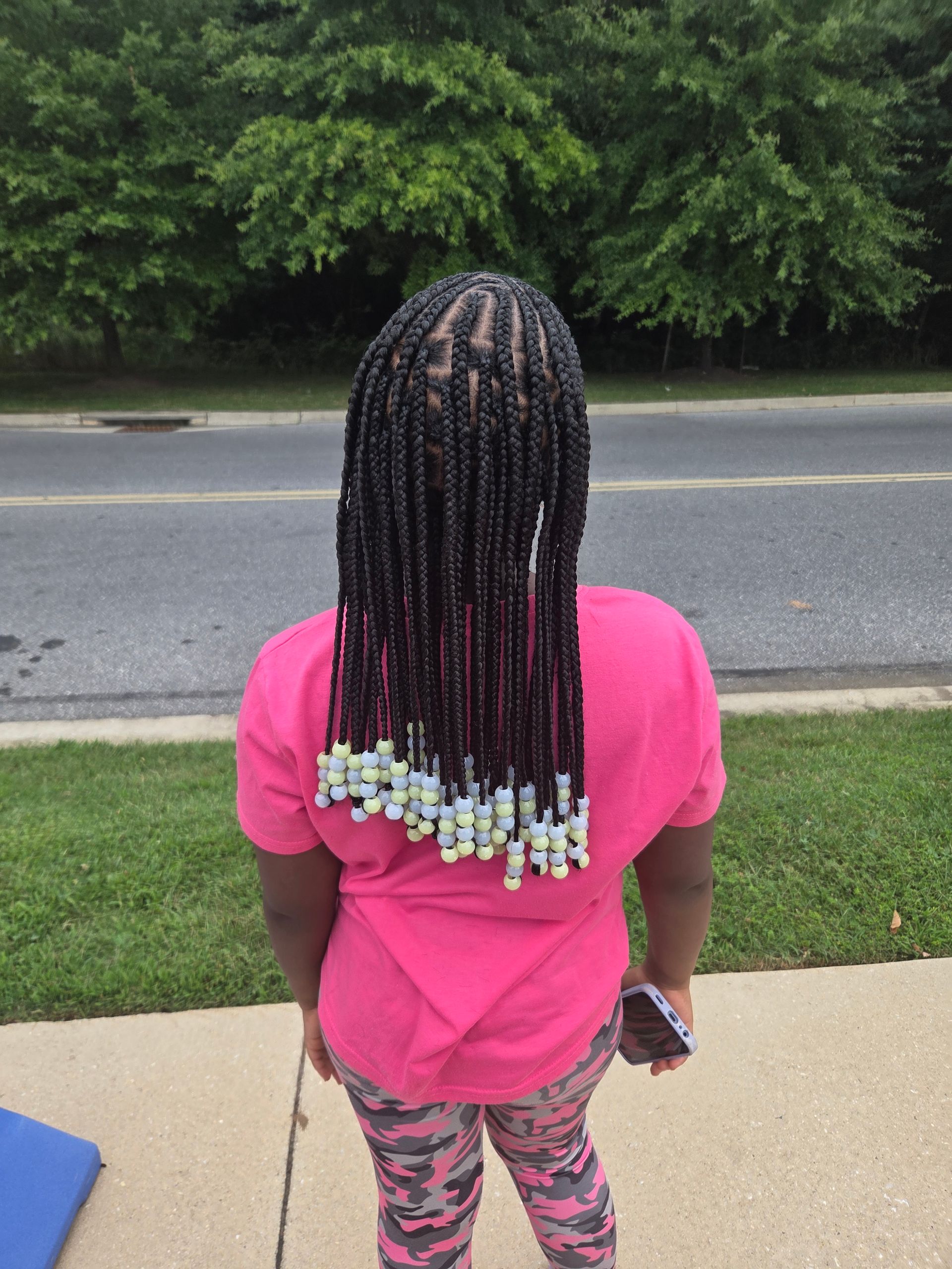 Girl with long black braided hair, wearing pink shirt and camouflage pants, standing outside. Beads are at the ends of the braids.