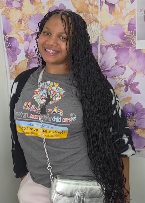 Woman with long micro braids smiles, wearing a grey t-shirt, silver purse, and cardigan. Background has floral pattern.