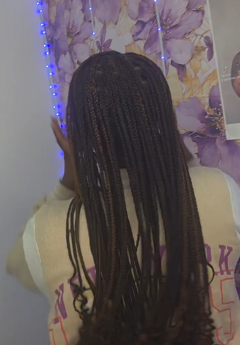 Woman with long braided hair, wearing a beige top, poses in front of a floral wall with blue string lights.