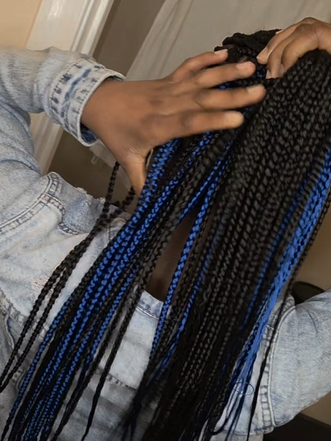 A person with black and blue box braids, wearing a denim jacket, touching their braids.