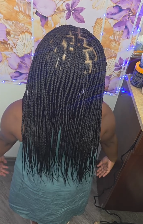 Black box braids, with decorative parting on the head, on a person wearing a teal top.