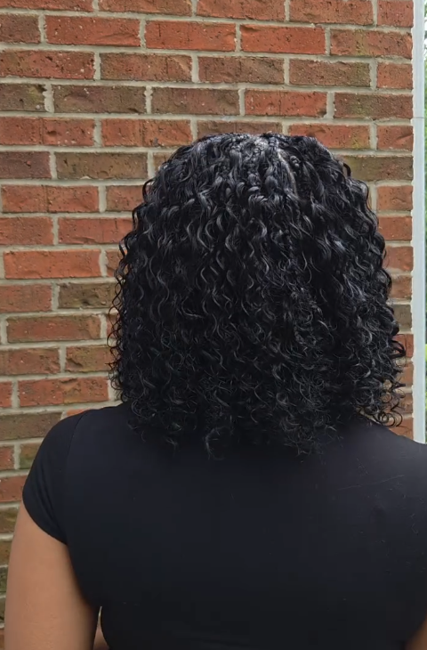 Woman with black, curly hair stands in front of a red brick wall. She wears a black shirt.