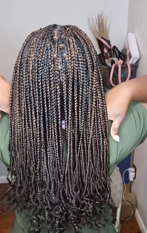 Woman with long box braids, brown and blonde highlights, green shirt, hands near her neck.