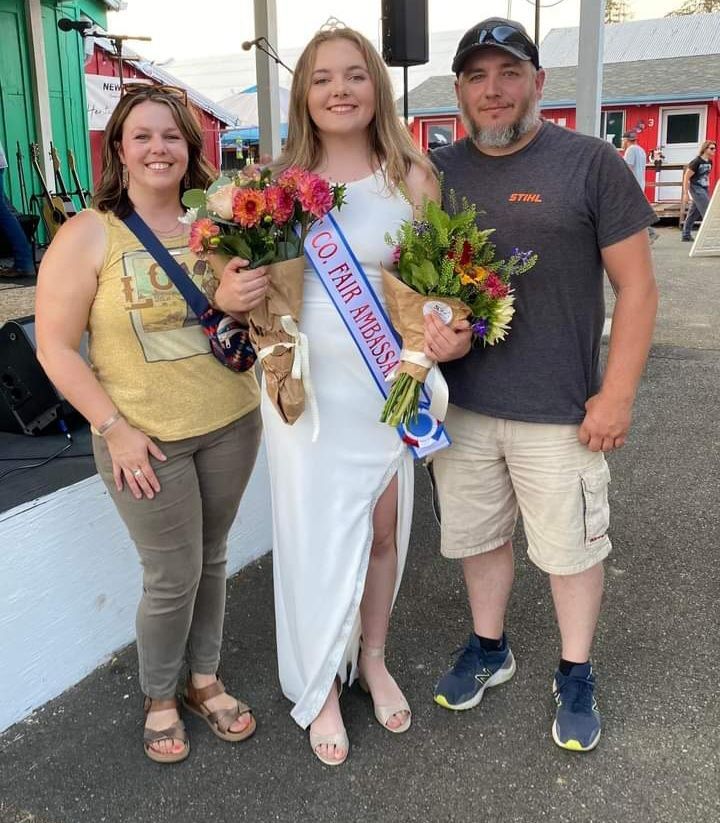 A woman in a white dress is holding flowers and a sash that says fair miss