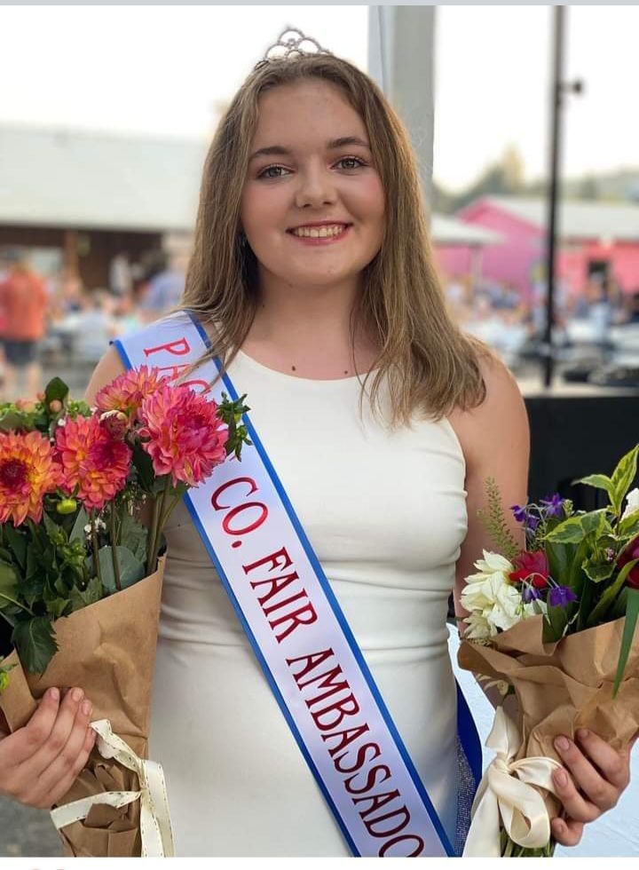 A woman wearing a sash that says co fair ambassador is holding flowers.
